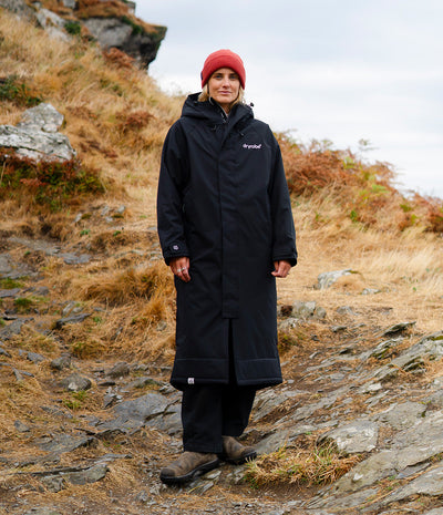 A person standing outdoors on a rocky hillside wearing a long black hooded waterproof jacket with a small white 'Troyjef' logo on the chest, a red beanie, dark pants, and brown boots.