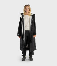 A woman stands against a light gray studio background wearing a long black hooded parka with a large white front panel and white zipper details, layered over a white top, black pants, and dark sneakers.