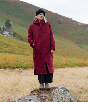 A woman wearing a long burgundy hooded raincoat, black trousers, and a black beanie stands on a rock in an outdoor mountain landscape.