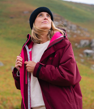 A woman wearing a burgundy Dryrobe hooded jacket with a bright pink inner lining, a cream sweatshirt, and a black beanie, standing outdoors against a blurred grassy hillside background.