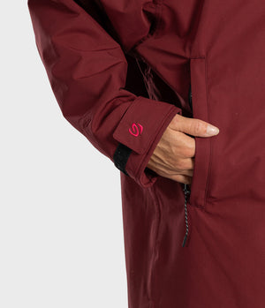 Close-up of a person wearing a dark red technical jacket, showing the sleeve cuff with a pink logo, a hand in a zippered pocket, and part of the torso against a light gray background.