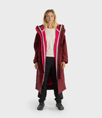 A woman modeling a long maroon hooded parka with a bright pink inner lining and white piping along the zipper, worn over a cream sweatshirt, black pants, and black sneakers against a light gray studio background.