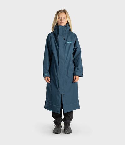A woman modelling a long, dark teal-blue hooded waterproof jacket with a small light blue 'dryrobe' logo on the left chest, paired with black pants and dark boots, against a light grey studio background.