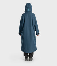 A person shown from the back wearing a long hooded waterproof coat in a muted blue-grey color, paired with black pants and dark shoes, against a plain light grey studio background.