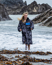 Woman running away from the sea smiling, wearing dryrobe Advance Long Sleeve REMIX Range change robe in Black Camo Black Blue