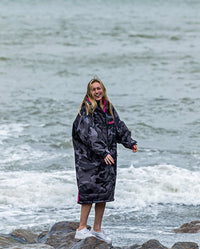 Woman stood on rocks next to the sea, wearing dryrobe Advance Long Sleeve REMIX Range change robe in Black Camo Pink Black