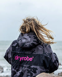 Woman stood on beach with back to camera, wearing dryrobe Advance Long Sleeve REMIX Range change robe in Black Camo Pink Black