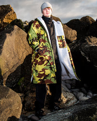 Man stood in front of rocks on beach, wearing dryrobe Advance Long Sleeve REMIX Range change robe in Camo Grey Green