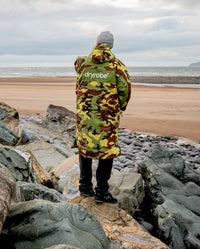 Man stood on rocks on beach with back to the camera, wearing dryrobe Advance Long Sleeve REMIX Range change robe in Camo Grey Green