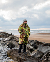 Man stood on beach in front of the sea, wearing dryrobe Advance Long Sleeve REMIX Range change robe in Camo Grey Green