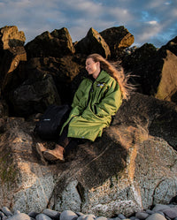 Woman sat on rocks on a beach, wearing dryrobe Advance Long Sleeve REMIX Range change robe in Forest Green Black Camo