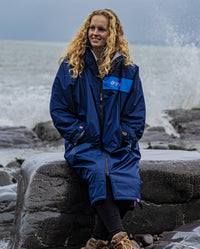 Woman sat on rock on a beach, wearing dryrobe Advance Long Sleeve REMIX Range change robe in Navy Grey Cobalt 