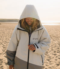 A woman wearing a Dryrobe hooded jacket stands on a sandy beach at sunset. The jacket is off-white on the upper body with gray lower panels and black zipper details.