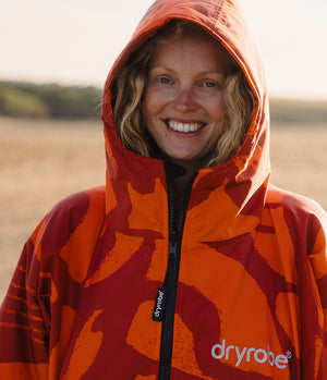A smiling woman wearing a hooded orange and red patterned Dryrobe jacket outdoors in warm sunlight.