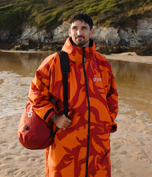 *MALE* A man standing on a sandy beach wearing a bright orange and red patterned Dryrobe waterproof jacket, with a red Dryrobe bag slung over his shoulder, against a rocky shoreline background.