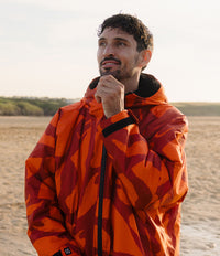 *MALE* A man wearing a bright orange and red camouflage hooded jacket, standing on a sandy beach with dunes in the background.