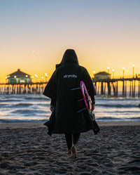 Woman carrying surfboard on Huntington Beach, wearing dryrobe® Lite changing robe