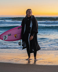 Woman stood in front of the ocean, carrying surfboard wearing dryrobe® Lite changing robe