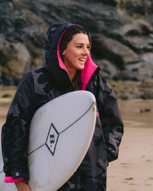 dryrobe¬Æ ambassador Lucy Campbell holding surfboard, wearing Black Camo Pink dryrobe¬Æ Advance Long Sleeve with hood up