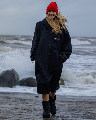 Woman walking on beach with sea behind her, wearing dryrobe Advance Long Sleeve changing robe in Black Red and Red dryrobe Beanie