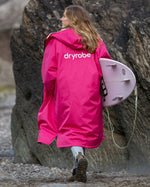 Woman walking along beach with back to the camera wearing dryrobe Advance long sleeve changing robe in Pink Light Grey