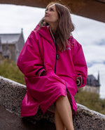 Woman sat on a wall looking up, wearing dryrobe Advance long sleeve changing robe in Pink Light Grey