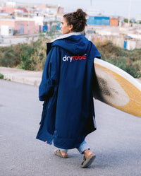 Woman walking along road carrying a yellow surfboard, wearing  Navy Grey dryrobe® Advance Long Sleeve