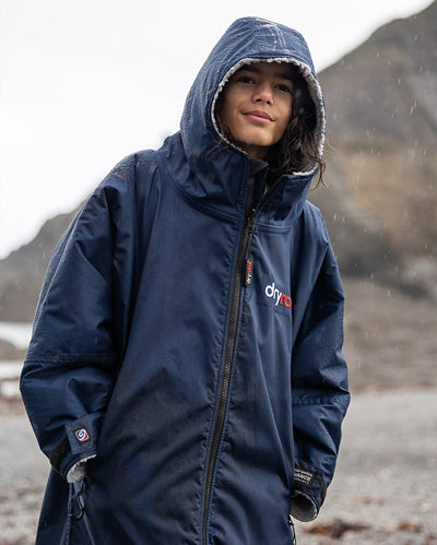 Boy stood in the rain on a beach wearing Navy Grey dryrobe® Advance Kids Long Sleeve with hood up 