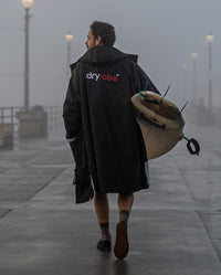 *MALE* carrying surfboard walking away from camera in the rain, wearing Black Grey dryrobe® Advance Long Sleeve