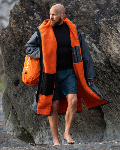 **MALE** Man stood in front of rocks on the beach, wearing dryrobe Advance Long Sleeve change robe in Dark Grey Orange, carrying compression bag 