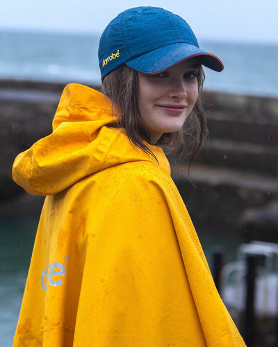 Woman stood in the rain on a beach, wearing Deep Sea Blue dryrobe Quick Dry Cap and Yellow Waterproof Poncho 