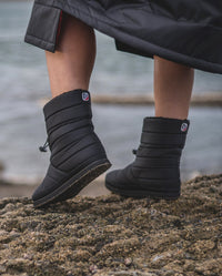 1|Close up of dryrobe® Eco Thermal Boots being worn on a beach, with one heel raised up